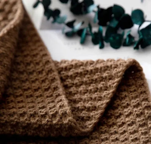 Plain beige knitted acrylic throw blanket with fringe on a grey sofa.