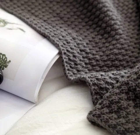 Plain beige knitted acrylic throw blanket with fringe on a grey sofa.