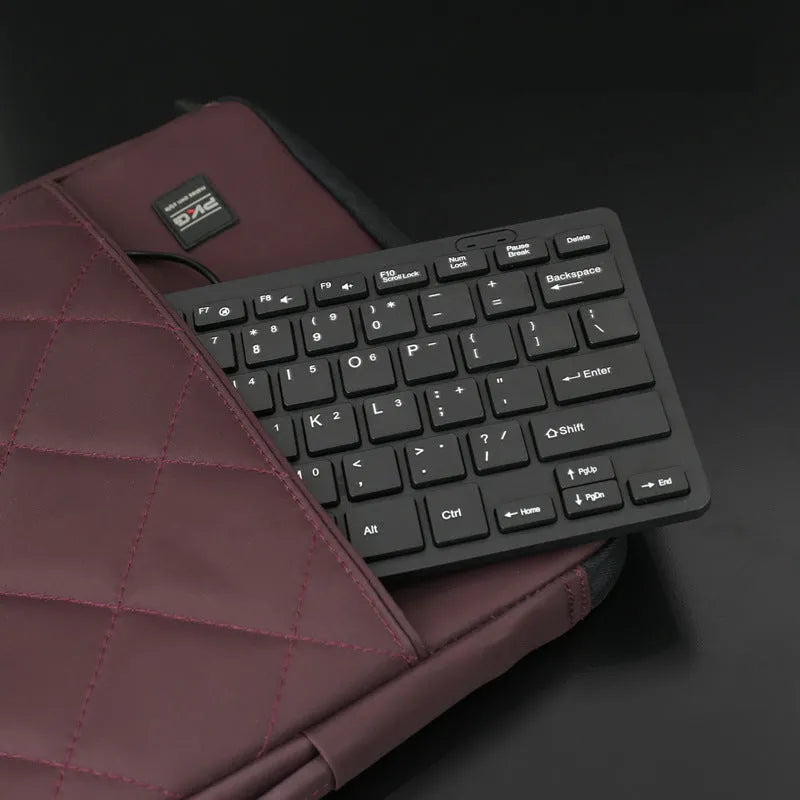 Compact wired USB keyboard in black, white, pink, or blue on a desk next to a laptop.