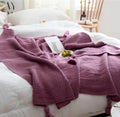 Plain beige knitted acrylic throw blanket with fringe on a grey sofa.