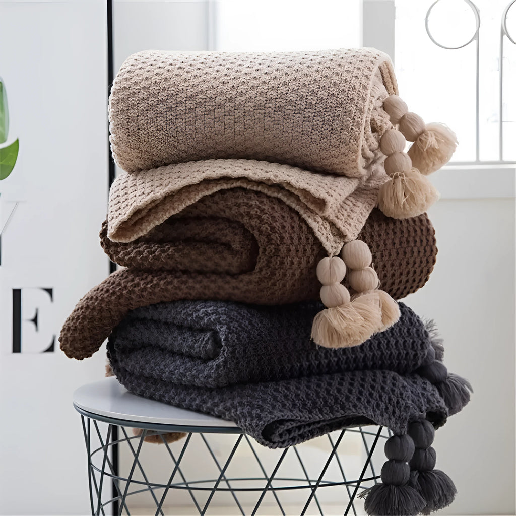 Plain beige knitted acrylic throw blanket with fringe on a grey sofa.
