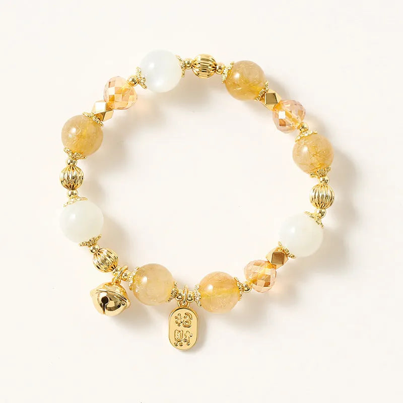 Gold-coloured rutilated quartz bracelet with a ringing bell charm on a wrist.