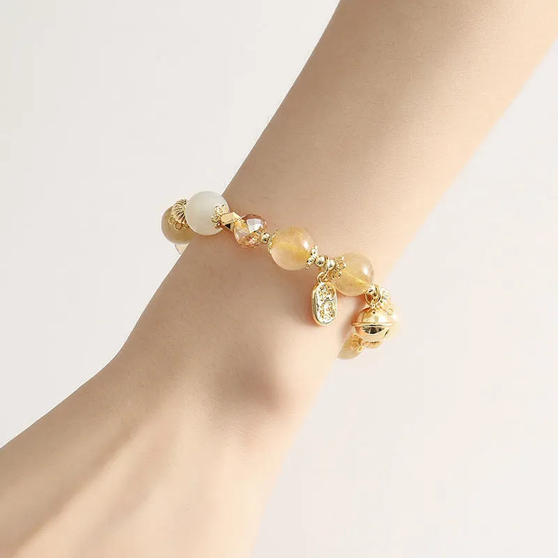 Gold-coloured rutilated quartz bracelet with a ringing bell charm on a wrist.
