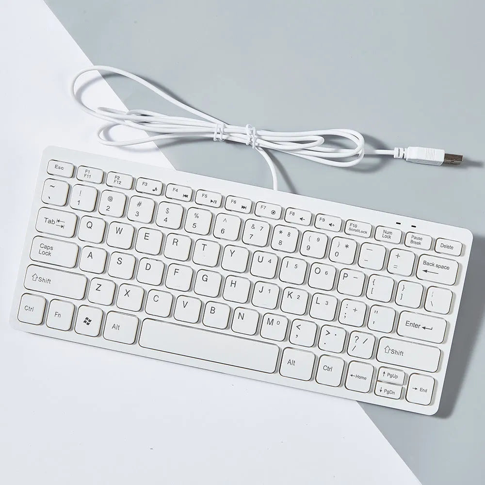 Compact wired USB keyboard in black, white, pink, or blue on a desk next to a laptop.